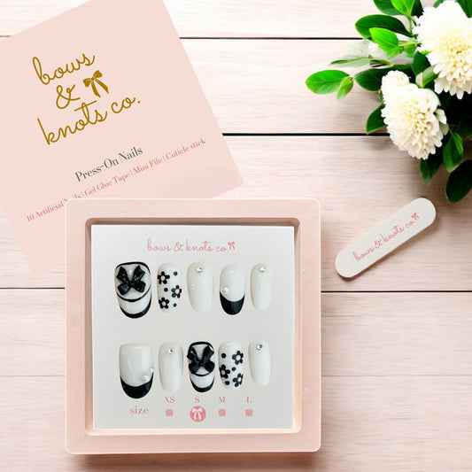 Set of press-on nails with a 'bows & knots co.' box on a wooden surface with flowers.