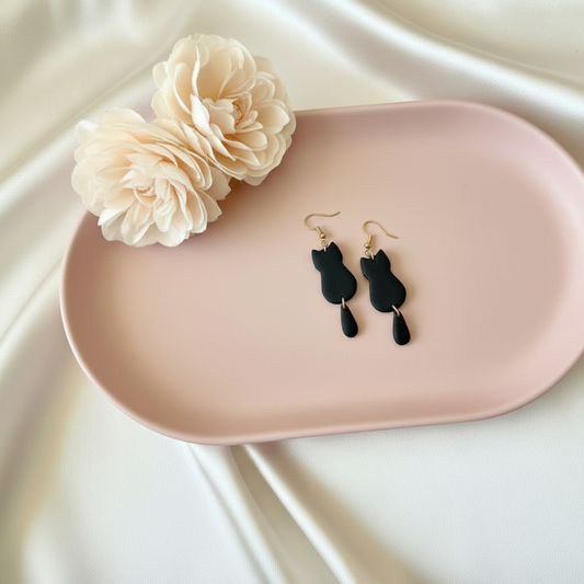Black cat-shaped earrings on a pink tray with a beige flower