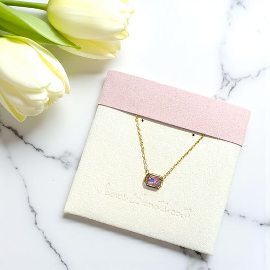 Gold necklace with a pink gemstone pendant on a white card with yellow tulips in the background.