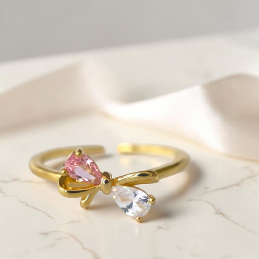 Gold bow-shaped ring with pink and clear gemstones on a light background