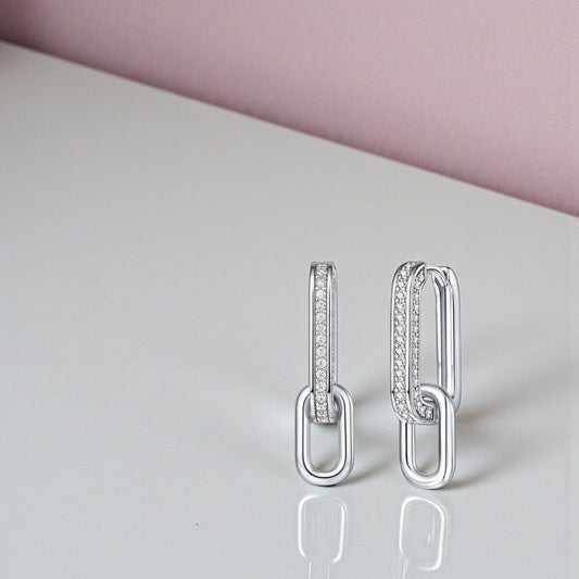 Pair of silver hoop earrings with embedded stones on a reflective surface with a pink background