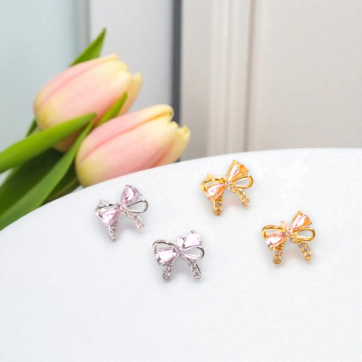 Bow-shaped earrings in gold and silver on a white surface with pink tulips in the background.