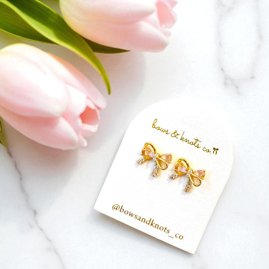 Gold bow-shaped earrings on a branded card with pink tulips in the background