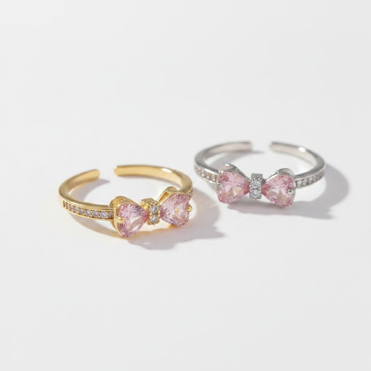 Two adjustable bow-shaped rings on a clean white background, one in yellow gold and one in silver, both featuring pink heart-shaped crystals and small clear accent stones.