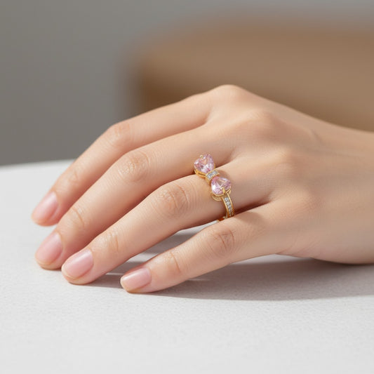 A close-up of a hand wearing a gold ring with two pink heart-shaped gemstones and small clear accent stones, resting on a light surface.