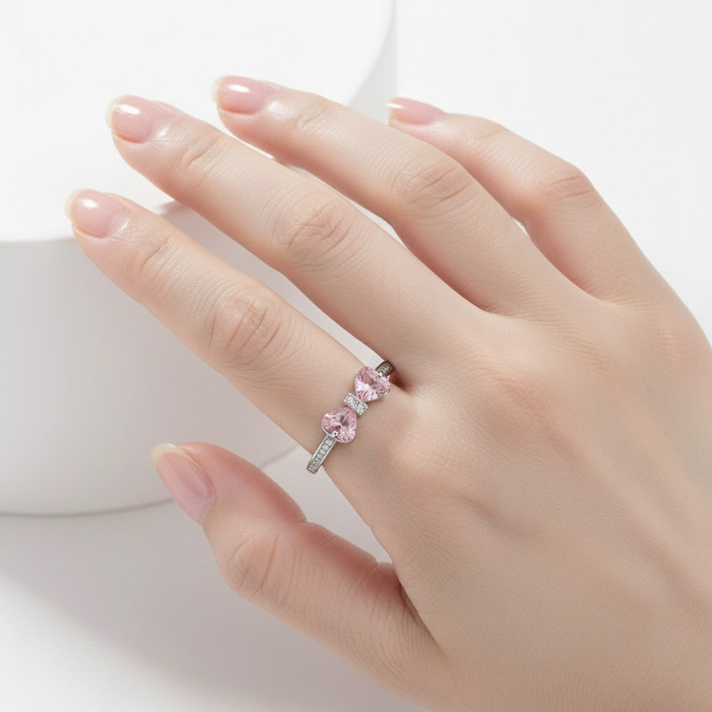 A close-up of a hand wearing a silver ring with three pink heart-shaped gemstones and small clear accent stones.