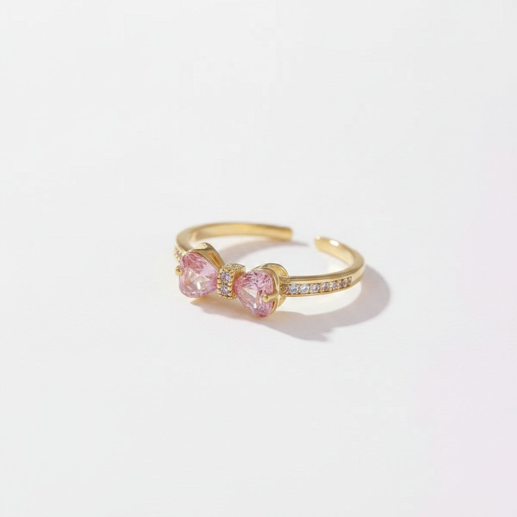 A gold adjustable ring featuring two heart-shaped pink gemstones in the center, accented by small clear stones along the band, set against a clean white background.