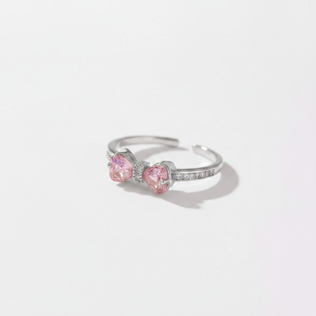 A silver-toned adjustable ring featuring two heart-shaped pink gemstones in a bow-like setting, accented with small clear stones along the band, photographed on a clean white background.