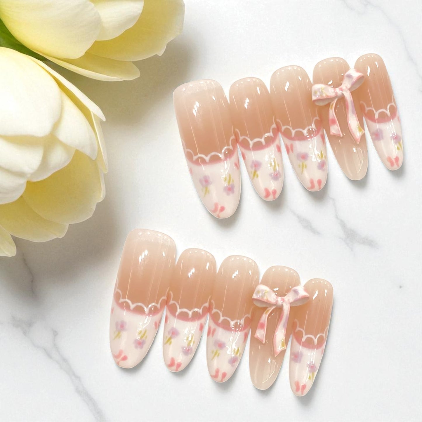 Decorative false nails with floral and bow designs on a marble surface with flowers.
