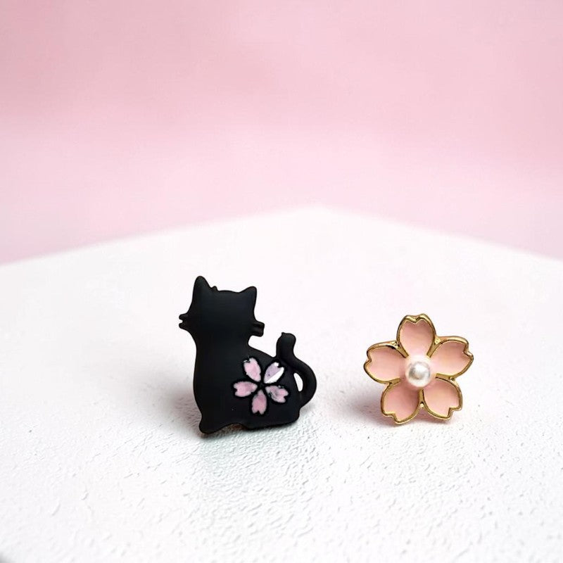Black cat-shaped earring with pink flower design and gold flower-shaped earring on a white surface.