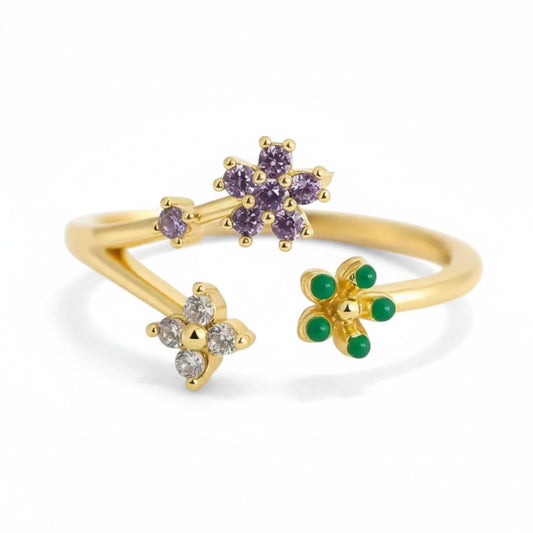 A gold open-band ring featuring three floral clusters: one with purple gemstones, one with clear stones, and one with green gemstones, shown on a white background.