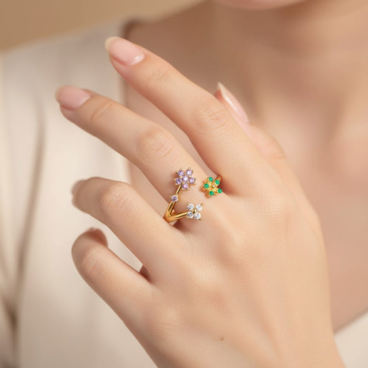A close-up of a hand wearing two delicate rings: one gold ring with small clear stones and a purple flower cluster, and one green flower ring with small clear stones.