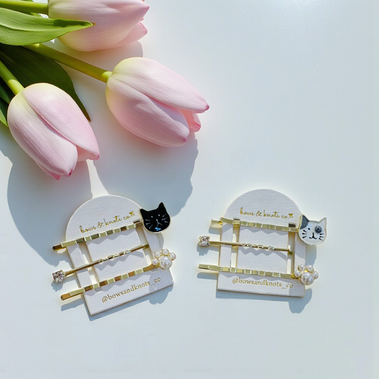 Two gold hair clips with cat designs on white cards, surrounded by pink tulips.