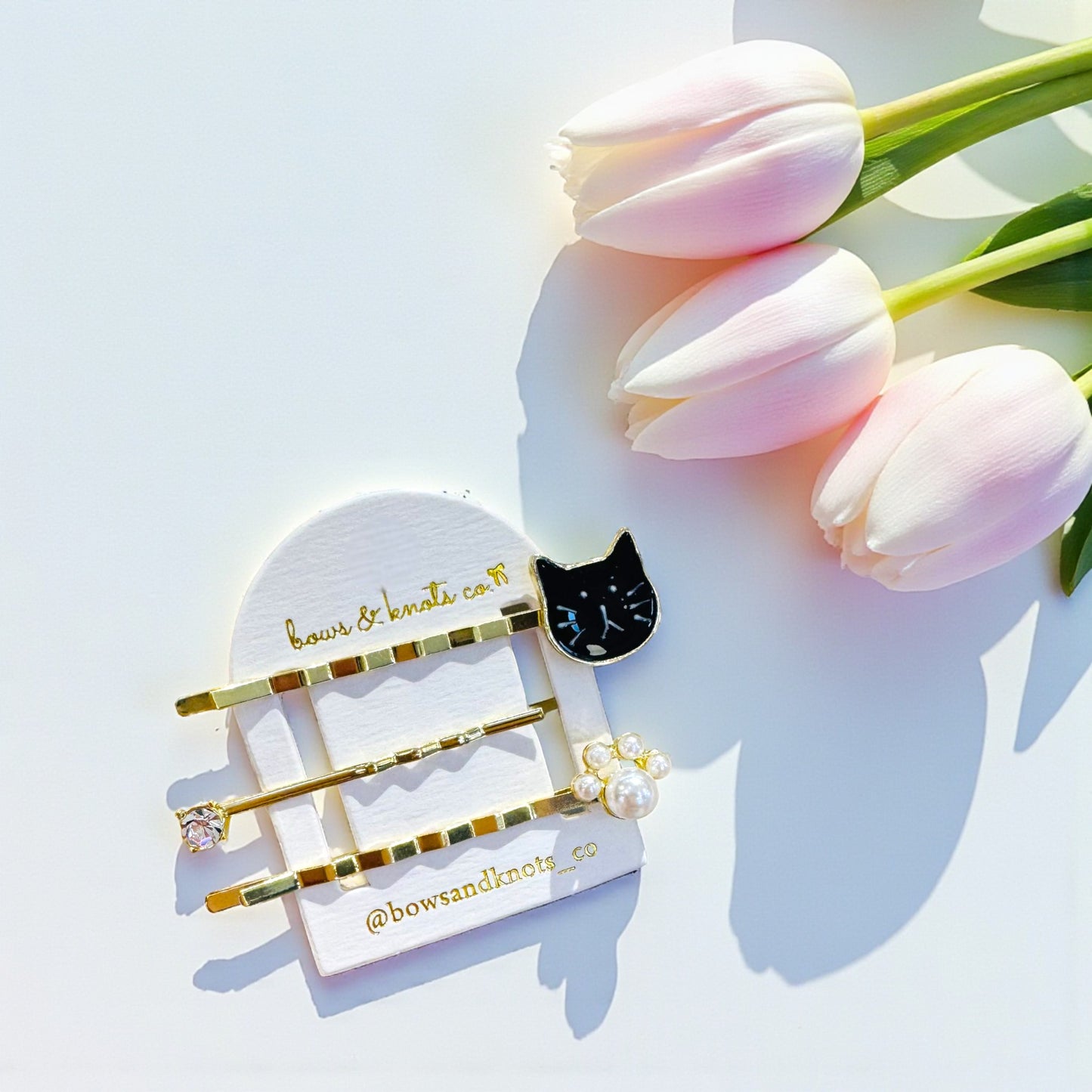 Set of hair clips with a black cat design on a card with 'bows & Knots' branding, next to pink tulips.