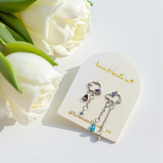 Pair of silver earrings with gemstones on a card with 'bows & knots co' branding, surrounded by white roses.