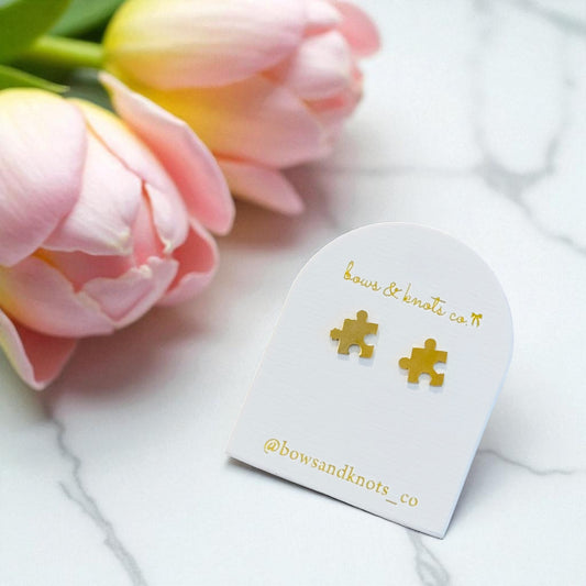 Gold puzzle piece earrings on a heart-shaped card with pink tulips in the background