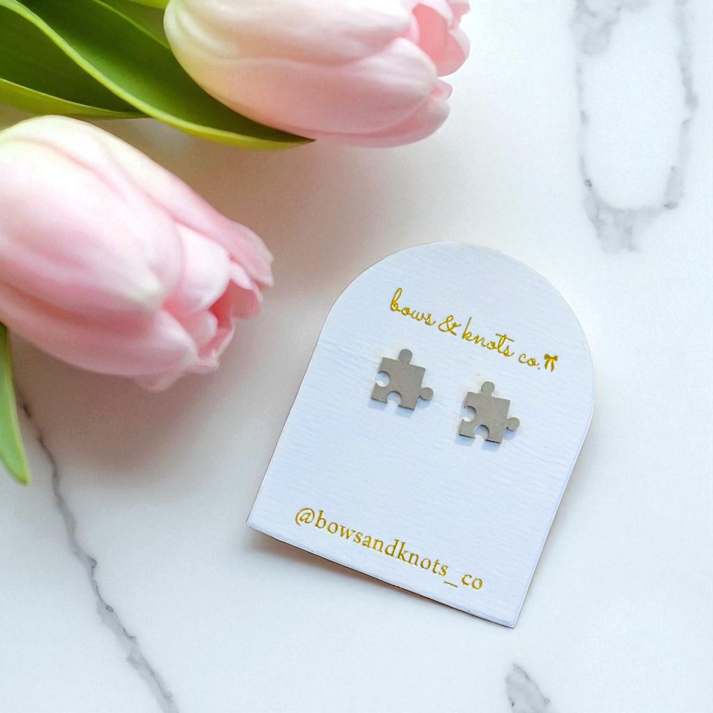 Pair of puzzle piece earrings on a 'bows & knots co' card with pink tulips in the background.
