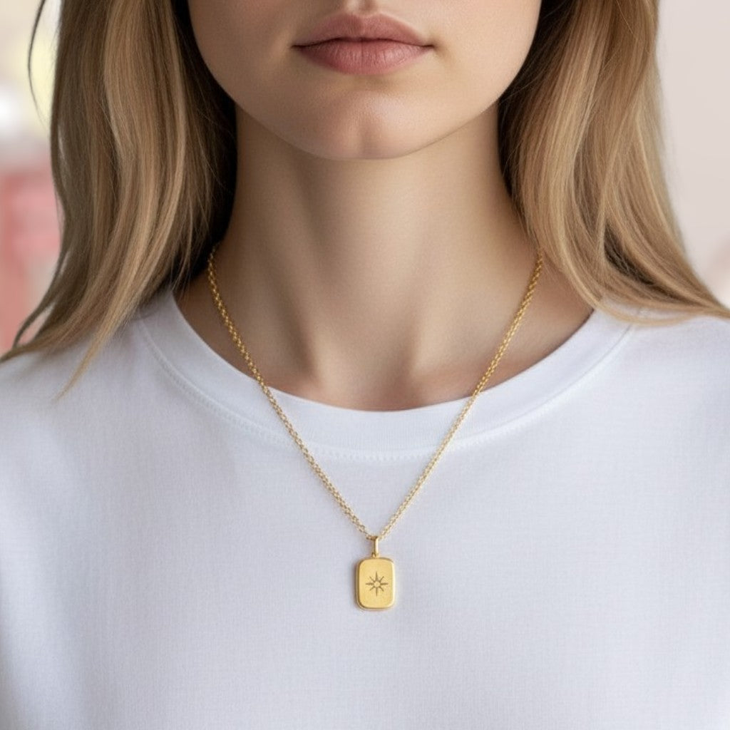 A close-up of a model wearing a delicate gold chain necklace with a small rectangular pendant featuring an engraved sunburst design, paired with a white crewneck top.