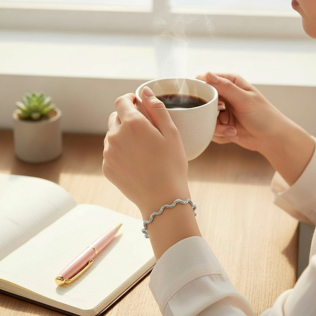 Person holding a steaming cup of coffee wearing a wavy chain bracelet from Bows and Knots Co. with a notebook and pen on a wooden table.