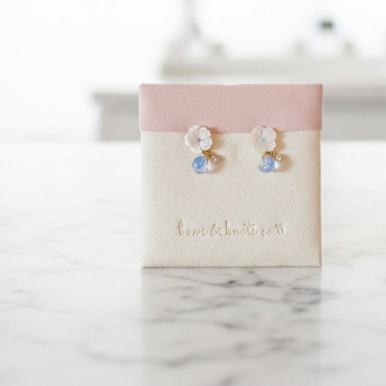 A pair of small floral stud earrings displayed on a cream jewelry card with a blush pink top band. The earrings feature white flower charms with clear faceted stones and small light blue drops, set on a white marble surface with a softly blurred background.